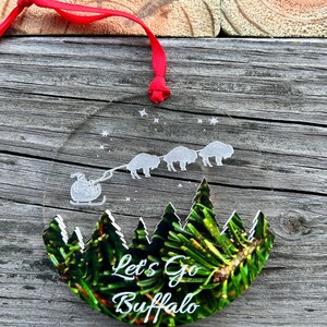 May include: A clear glass ornament with a green pine tree design and the text "Let's Go Buffalo". The ornament is decorated with white stars and a silhouette of a sleigh pulled by four buffalo.
