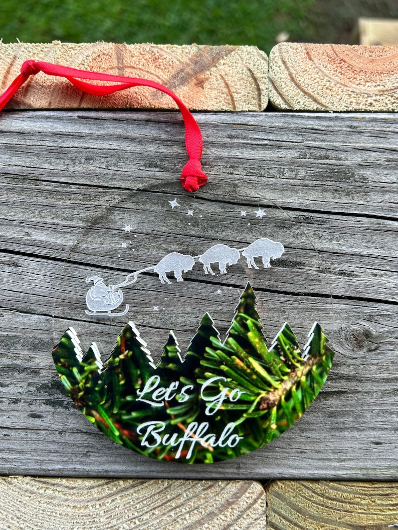 May include: A clear glass ornament with a green pine tree design and the text "Let's Go Buffalo". The ornament is decorated with white stars and a silhouette of a sleigh pulled by four buffalo.