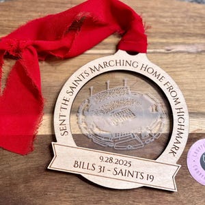 May include: A wooden ornament with a stadium design and the text "SENT THE SAINTS MARCHING HOME FROM HIGHMARK". It includes the date "9.28.2025" and the score "BILLS 31 - SAINTS 19". A red ribbon is attached for hanging.
