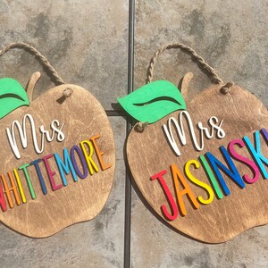 May include: Two wooden apple-shaped signs with a green leaf on top. The signs are painted brown and have the text "Mrs. Whittemore" and "Mrs. Jasinski" in rainbow letters.