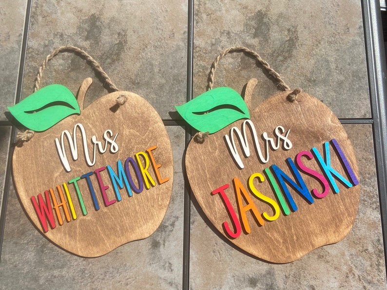 May include: Two wooden apple-shaped signs with a green leaf on top. The signs are painted brown and have the text "Mrs. Whittemore" and "Mrs. Jasinski" in rainbow letters.