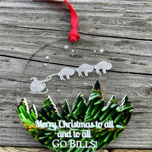 May include: Round, clear Christmas ornament with a green pine tree background. The ornament features a white silhouette of Santa Claus pulling a sleigh with four buffalo. The text "Merry Christmas to all and to all GO BILLS!" is written in white.