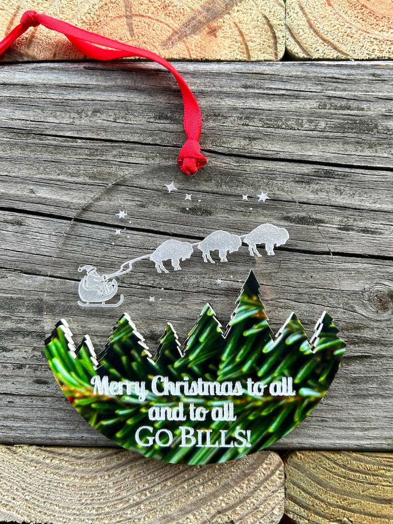 May include: Round, clear Christmas ornament with a green pine tree background. The ornament features a white silhouette of Santa Claus pulling a sleigh with four buffalo. The text "Merry Christmas to all and to all GO BILLS!" is written in white.