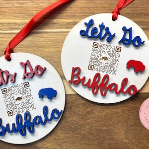 May include: Two white round wooden ornaments with the text "Let's Do Buffalo" in red and blue letters. Each ornament has a black and white QR code and a blue or red buffalo silhouette.