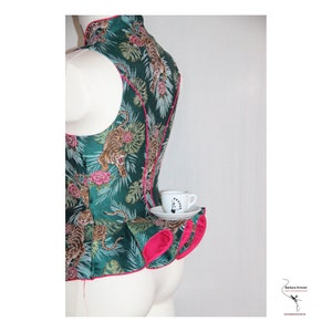 Może przedstawiać: Zielony top bez rękaw&oacute;w z r&oacute;żową lam&oacute;wką i nadrukiem tygrysa. Top ma falbanastą dolną część i mały biały kubek do herbaty z podstawką leżą na falbanie.