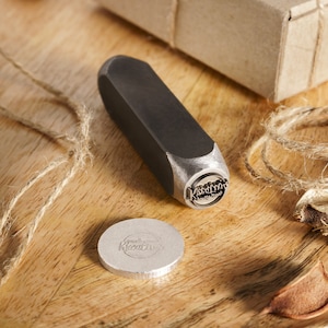 May include: A close-up of a metal stamp with the words "Kustom Kreations" engraved on it. The stamp is on a wooden surface next to a small, round, silver metal disc with the same words engraved on it. A brown box with twine and a red wax seal is in the background.