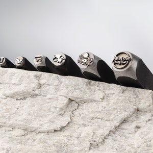 May include: Six metal stamps with different designs, including a bird, a cow, a dragon, a tree, and the words "Roadhouse Country Grill". The stamps are arranged in a row on a light gray textured surface.