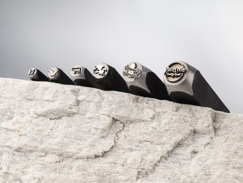 May include: Six metal stamps with different designs, including a bird, a cow, a dragon, a tree, and the words "Roadhouse Country Grill". The stamps are arranged in a row on a light gray textured surface.