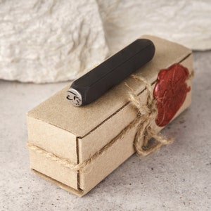 May include: A black metal stamp with a design of a heart with an arrow through it, packaged in a brown cardboard box with twine and a red wax seal.