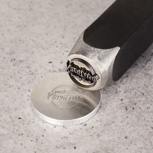 May include: A silver metal stamp with the text "Good Things Grow Here" and a black handle. The stamp is being used to create an impression on a silver metal disc with the text "Good Things Grow Here" on it.
