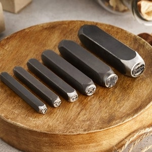 May include: A set of six metal stamping tools with different designs on a wooden surface. The tools are used for creating custom jewelry and other crafts.