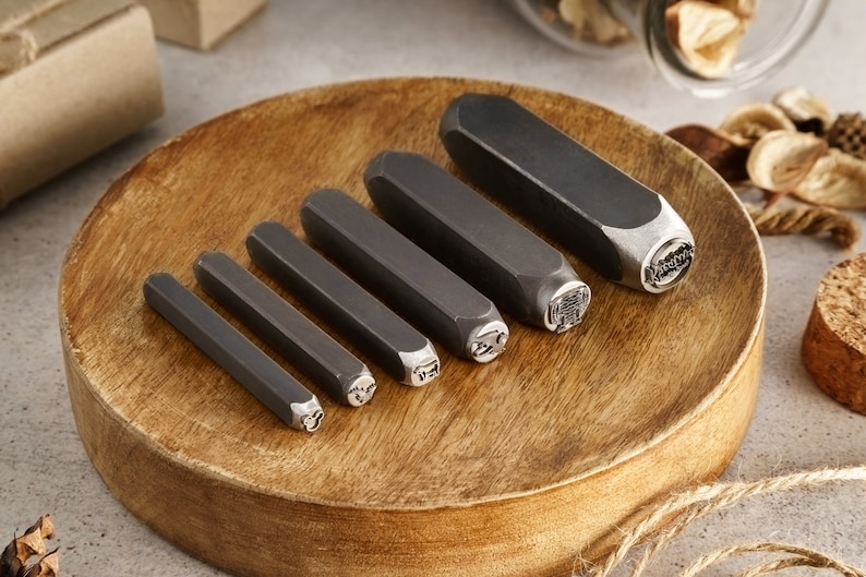 May include: A set of six metal stamping tools with different designs on a wooden surface. The tools are used for creating custom jewelry and other crafts.