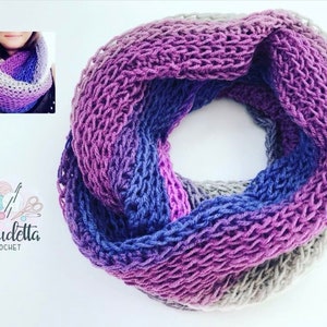 Op de afbeelding: Een handgemaakte infinity sjaal in tinten paars, blauw en grijs. De gebreide sjaal heeft een gradiënteffect, dat overgaat van lichte naar donkere kleuren. De sjaal wordt gepresenteerd op een wit oppervlak. Het Claudetta Crochet-logo staat linksonder.
