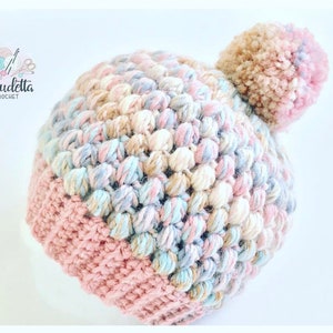 Könnte beinhalten: Handgefertigte, pastellfarbene Häkelmütze mit strukturiertem Noppenstich-Design. Die Mütze hat einen rosa Rand und einen flauschigen Bommel oben. Das Logo "Claudetta Crochet" ist links oben sichtbar.