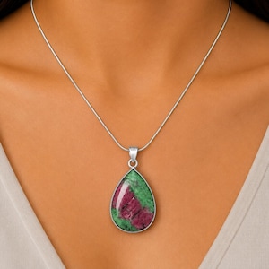 Può includere: Una collana con pendente a forma di goccia caratterizzata da una gemma di rubino e zoisite lucida con inclusioni verde brillante e rosso magenta intenso, incastonata in una semplice montatura in metallo argentato e appesa a una sottile catena a serpente argentata.