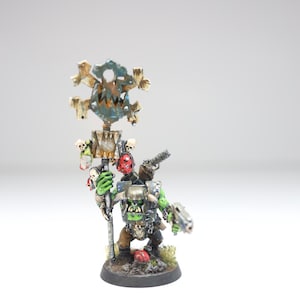 May include: A green and brown painted miniature figurine of an Orc holding a weapon and a banner with a skull and crossbones design. The banner is decorated with skulls and red and gold accents.
