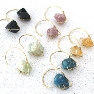 May include: A selection of gemstone earrings. Each earring has a raw, irregularly shaped gemstone in various colours, including black, pink, yellow, green, and blue, suspended from a gold-coloured wire hook.