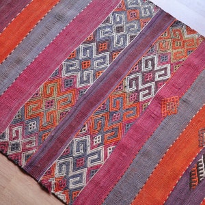 May include: A vibrant rug featuring diagonal stripes in orange, purple, and grey. Some stripes display detailed geometric patterns in white, orange, and grey. The rug's woven texture suggests a handmade or traditional design, adding a touch of warmth.