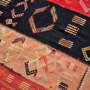 Puede incluir: Un primer plano de una alfombra tejida colorida con patrones geométricos en tonos de rojo, naranja, amarillo, azul y blanco. La alfombra está hecha de lana y tiene un diseño tradicional.