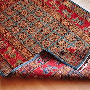 May include: A rectangular, hand-woven rug with a detailed pattern. The rug features a central teal field with repeating square motifs, framed by a red border with animal and geometric designs. The rug is partially rolled up, revealing its underside.