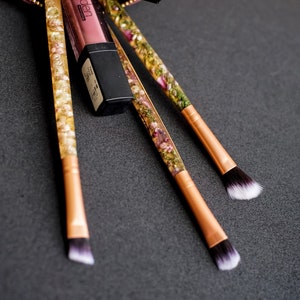 May include: Makeup brushes with clear handles filled with dried flowers and gold-colored metal ferrules. A tube of lipstick and a black eyeshadow palette are also present. The items are arranged on a dark gray surface.