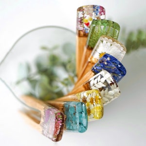 May include: A set of seven wooden hair sticks with colorful resin tops. Each top features pressed flowers and botanicals, including ferns, daisies, and blue flowers. The sticks are arranged in a fan shape, with the resin tops facing the viewer.