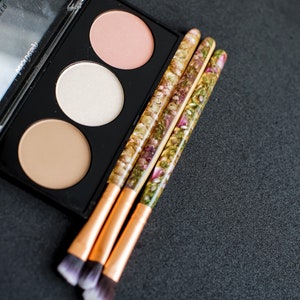 May include: A black makeup palette with three circular pans of blush, highlighter, and bronzer. Three makeup brushes with gold ferrules and handles filled with dried flowers are next to the palette. The brush tips are white and dark brown.