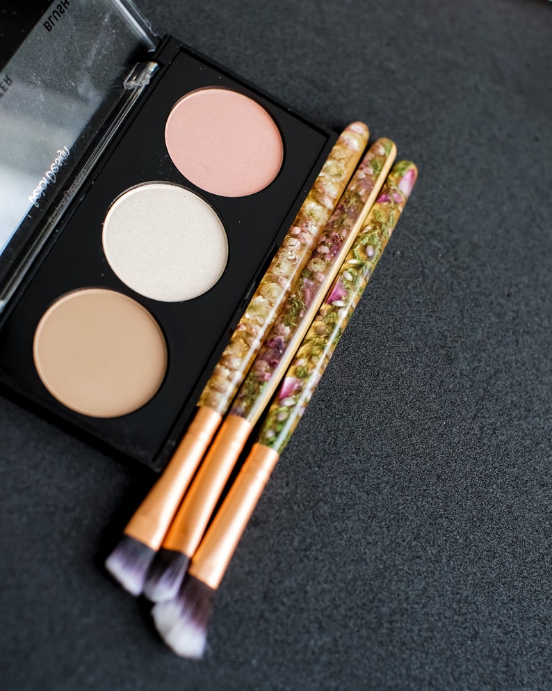 May include: A black makeup palette with three circular pans of blush, highlighter, and bronzer. Three makeup brushes with gold ferrules and handles filled with dried flowers are next to the palette. The brush tips are white and dark brown.