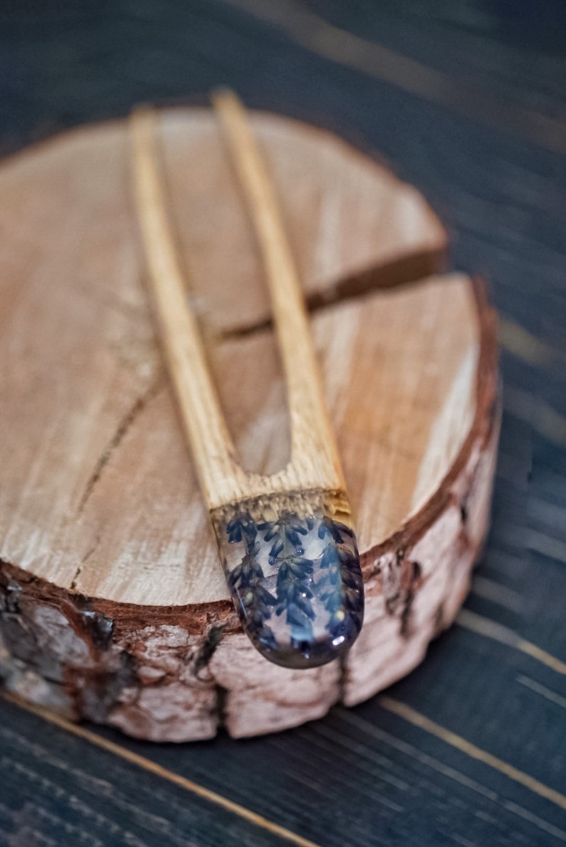 May include: A wooden hair fork with a clear resin tip containing pressed lavender flowers. The fork is resting on a wooden surface.
