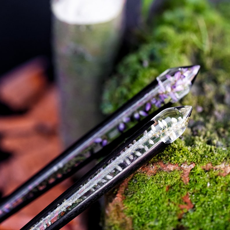 May include: Two black hair chopsticks with clear resin tips. One chopstick has purple flowers embedded in the resin, the other has white flowers. The chopsticks are resting on a bed of green moss.