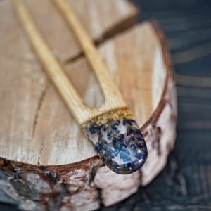 May include: A wooden hair fork with a clear resin tip containing pressed purple flowers. The fork is resting on a wooden surface.