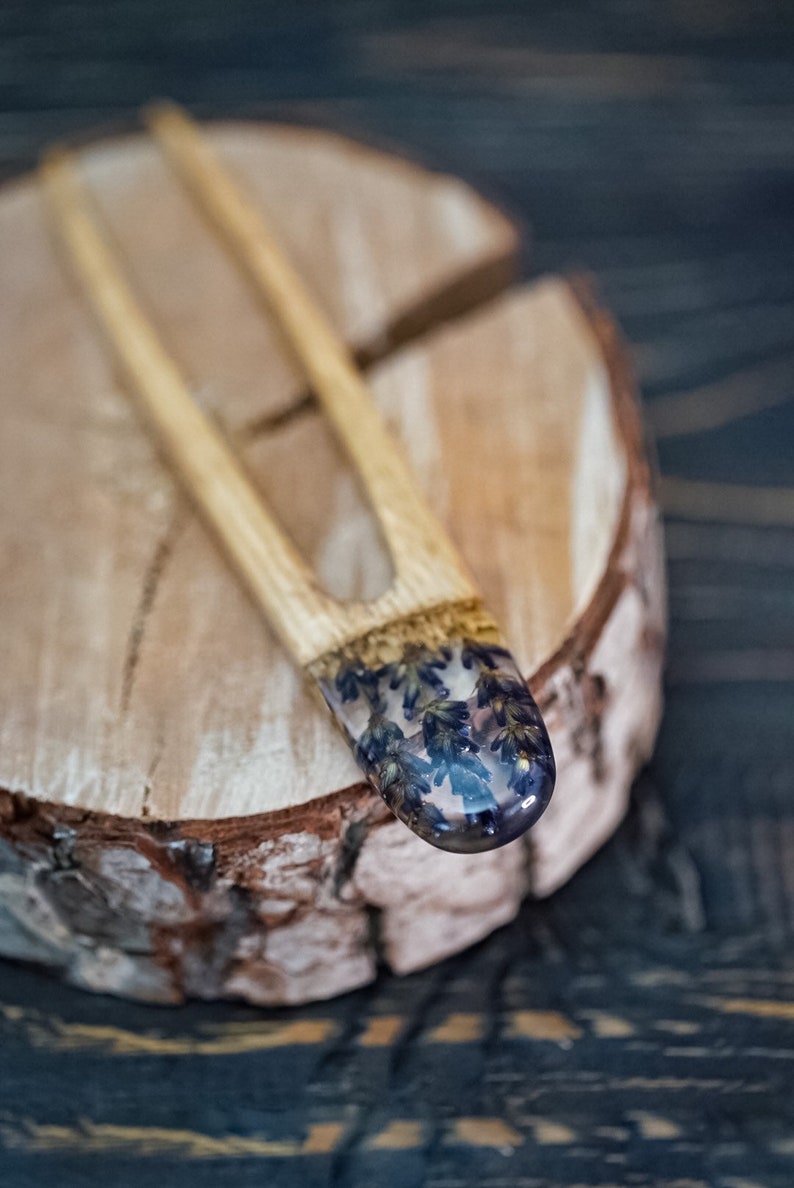 May include: A wooden hair fork with a clear resin tip containing pressed purple flowers. The fork is resting on a wooden surface.