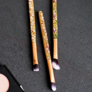 May include: Three makeup brushes with wooden handles and floral resin accents. The brush heads are white and black. The handles have a gold-colored ferrule. The brushes are arranged on a dark gray surface, with a makeup palette in the corner.