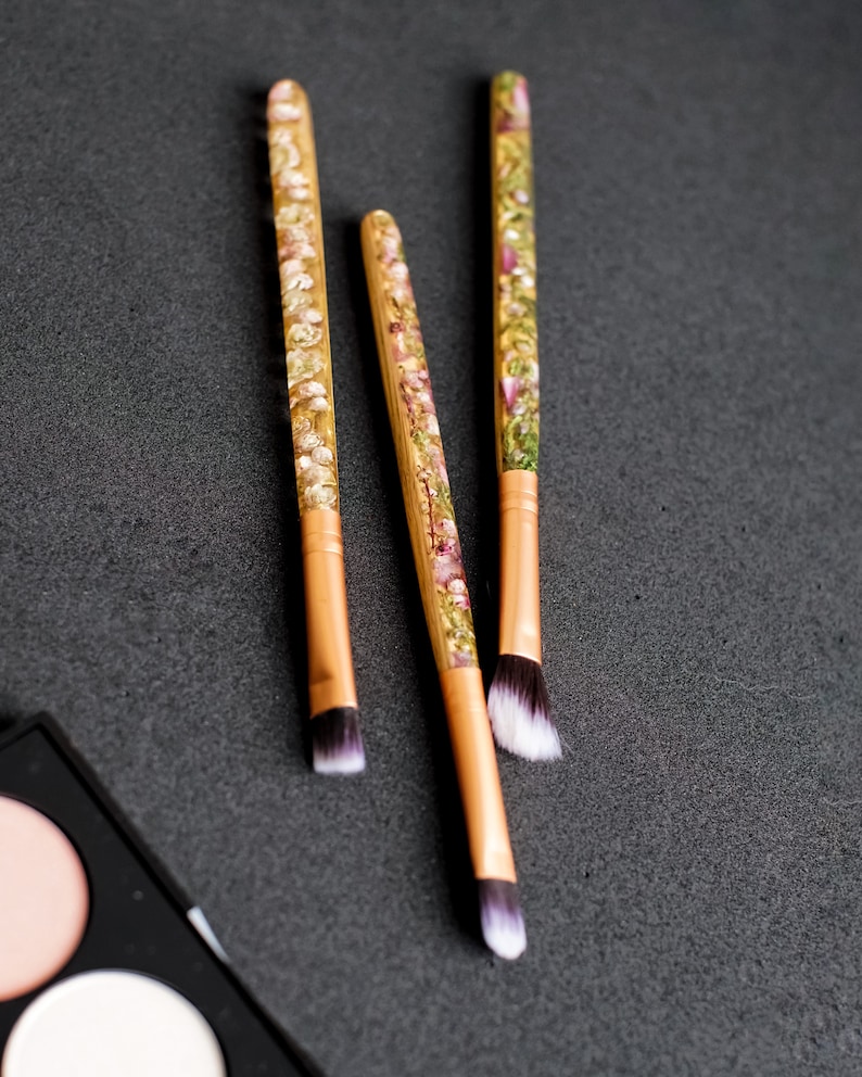 May include: Three makeup brushes with wooden handles and floral resin accents. The brush heads are white and black. The handles have a gold-colored ferrule. The brushes are arranged on a dark gray surface, with a makeup palette in the corner.