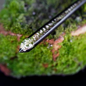 May include: A black and silver hair stick with a clear resin section containing white flecks. The stick is resting on a bed of green moss.