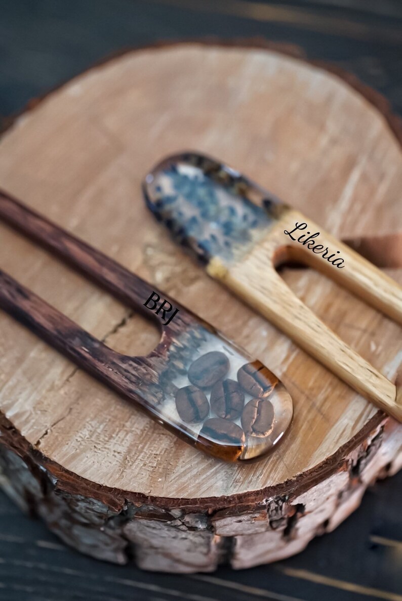 May include: Two wooden hair sticks with resin accents. One stick has a clear resin section with coffee beans embedded inside. The other stick has a blue and white resin section. The sticks are on a wooden surface. The stick with the coffee beans has the text "BRJ" on it. The stick with the blue and white resin has the text "Likeria" on it.