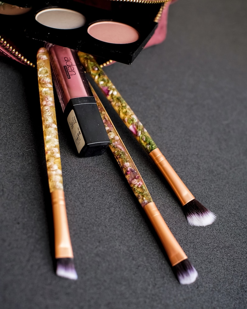 May include: Makeup brushes with clear handles filled with dried flowers and gold-colored metal ferrules. A tube of lipstick and a black eyeshadow palette are also present. The items are arranged on a dark gray surface.