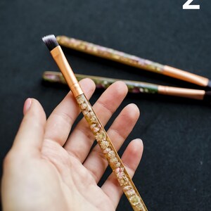 May include: A makeup brush with a gold-colored metal ferrule and a handle filled with small, dried flowers. The brush has a flat, angled head with black and white bristles. Two other brushes are visible in the background.