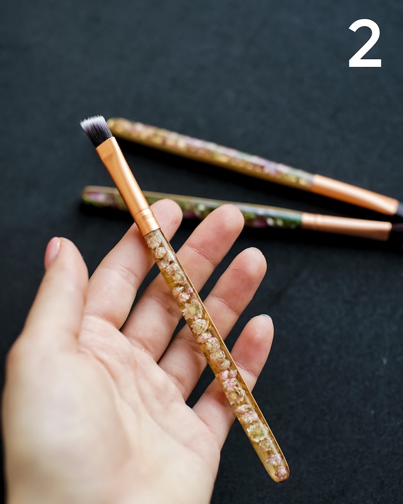 May include: A makeup brush with a gold-colored metal ferrule and a handle filled with small, dried flowers. The brush has a flat, angled head with black and white bristles. Two other brushes are visible in the background.