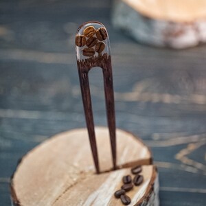 May include: A wooden hair fork with a clear resin section filled with coffee beans. The fork is standing upright on a wooden base.