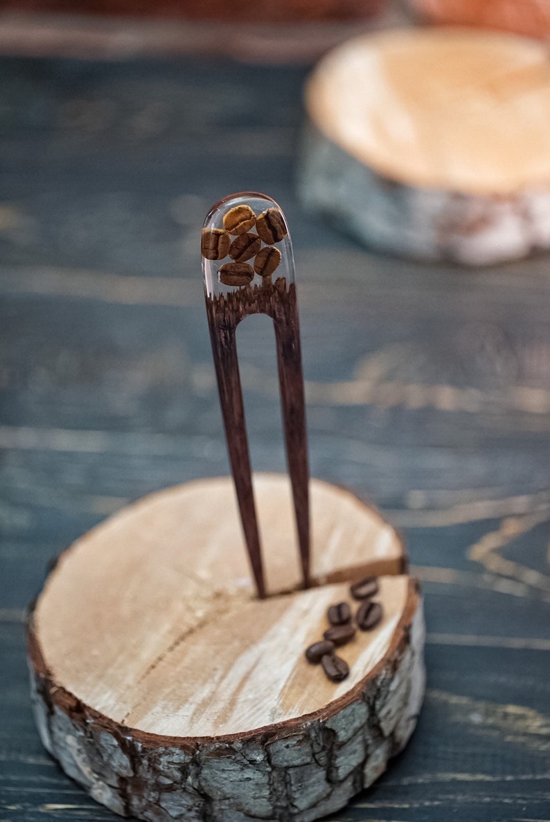 May include: A wooden hair fork with a clear resin section filled with coffee beans. The fork is standing upright on a wooden base.