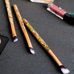 May include: Three makeup brushes with light brown handles and white and black bristles. The handles are decorated with a clear resin filled with dried flowers and gold flakes. Other makeup items are in the background.