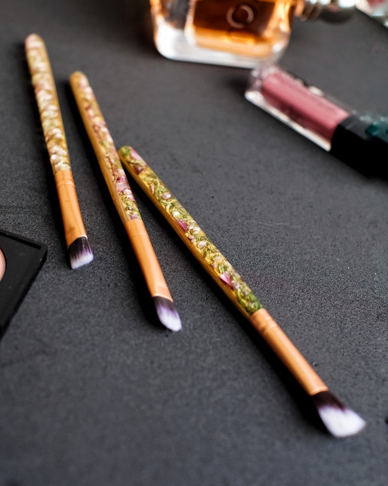 May include: Three makeup brushes with light brown handles and white and black bristles. The handles are decorated with a clear resin filled with dried flowers and gold flakes. Other makeup items are in the background.