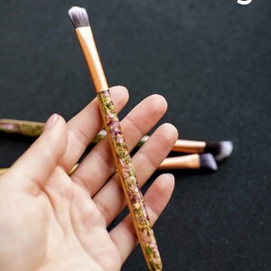 May include: A makeup brush with a copper-colored ferrule and a handle filled with dried flowers. The brush has a flat, angled head with white and gray bristles. The number "3" is in the upper right corner.