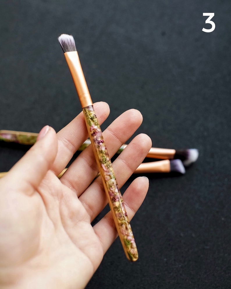 May include: A makeup brush with a copper-colored ferrule and a handle filled with dried flowers. The brush has a flat, angled head with white and gray bristles. The number "3" is in the upper right corner.