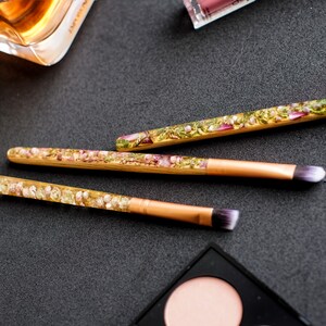 May include: Three makeup brushes with clear handles filled with dried flowers and gold-colored metal ferrules. The brush heads are white and purple. The brushes are on a dark surface with other makeup items.