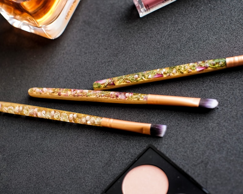 May include: Three makeup brushes with clear handles filled with dried flowers and gold-colored metal ferrules. The brush heads are white and purple. The brushes are on a dark surface with other makeup items.