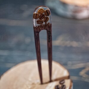 May include: A wooden hair fork with a clear resin top containing coffee beans. The fork is engraved with the word "Milania".
