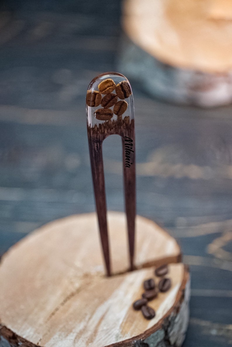 May include: A wooden hair fork with a clear resin top containing coffee beans. The fork is engraved with the word "Milania".