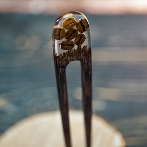 May include: A wooden hair fork with a clear resin top containing coffee beans. The fork is resting on a wooden surface.